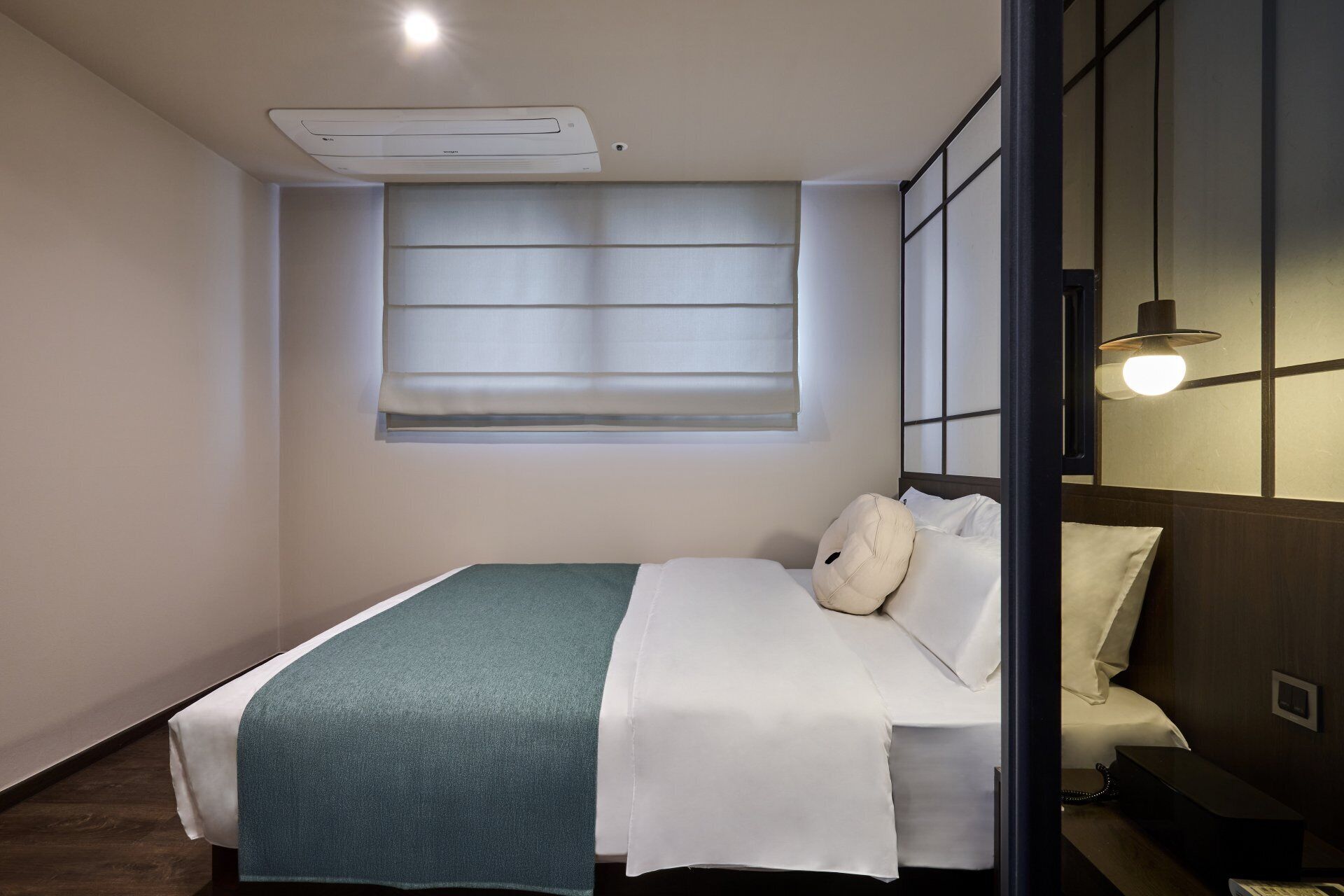 A hotel room with a bed, a window with blinds, a bedside table, and a wall-mounted light fixture.