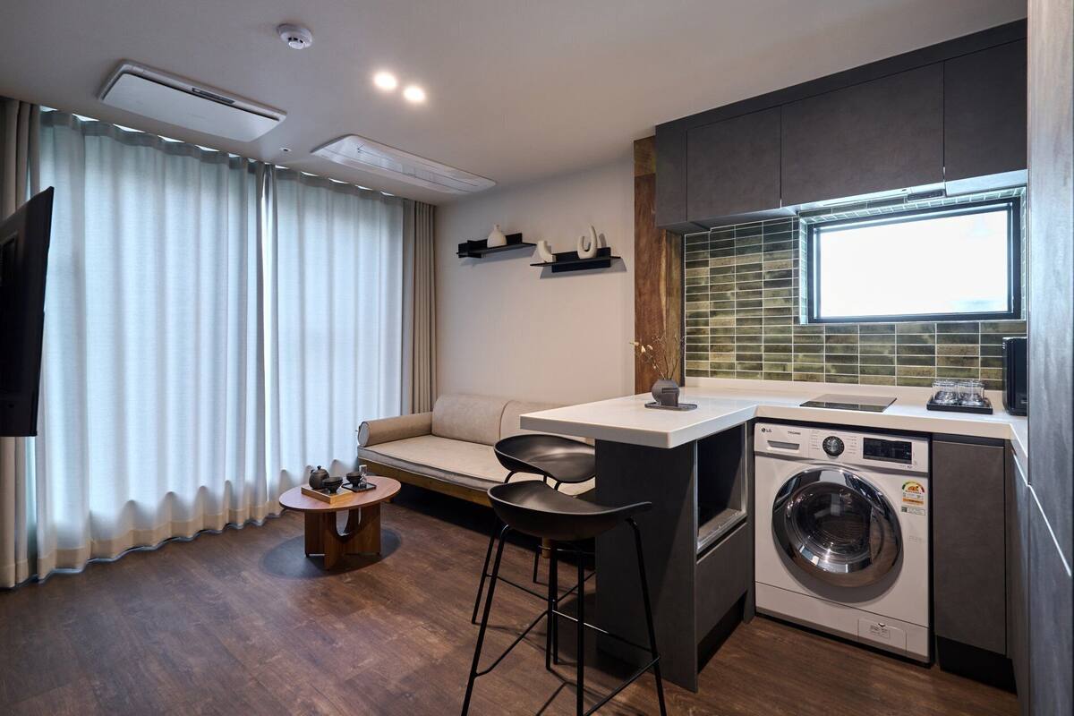 Une cuisine compacte équipée d’un lave-linge, un coin repas avec un canapé et des tabourets, et un téléviseur fixé au mur.
