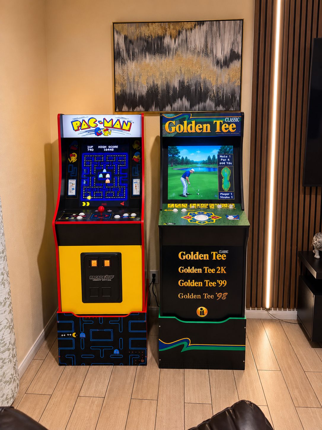 Deux bornes d’arcade, l’une portant l’inscription « PAC-MAN » et l’autre « Golden Tee », se trouvent dans une pièce dont le sol est en bois et dont un des murs est orné d’un tableau.