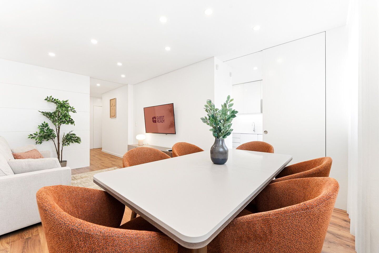 Ein modernes Hotelzimmer mit einem weißen Tisch, orangefarbenen Stühlen, einer Topfpflanze und einem Schild mit der Aufschrift 'GUEST READY'.