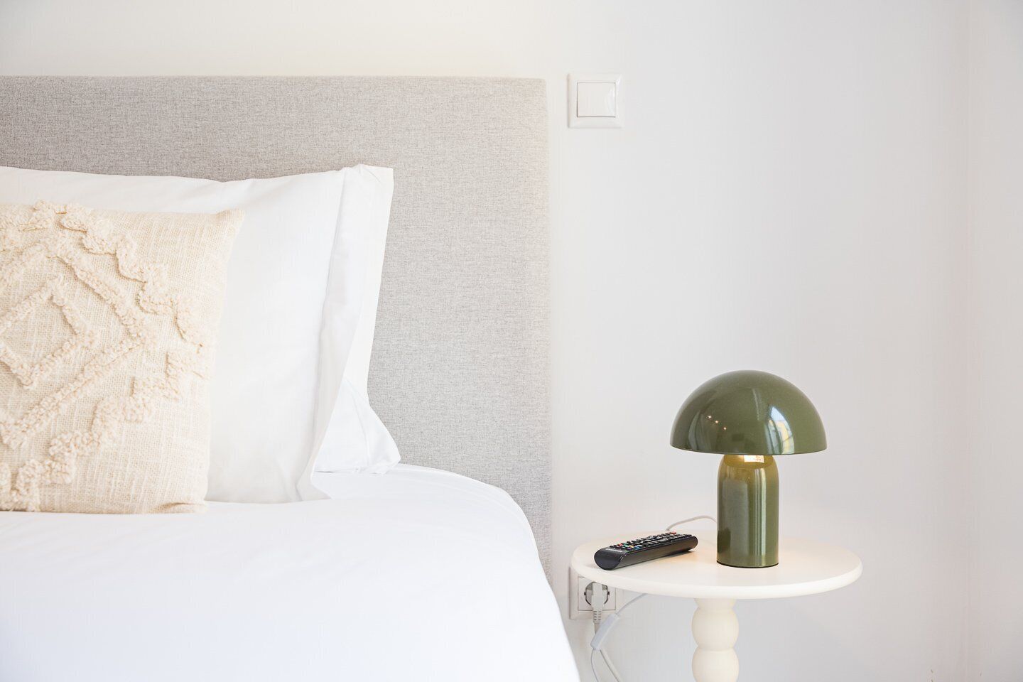 A neatly made bed with a white and beige pillow, a green lamp on a white side table, and a remote control.