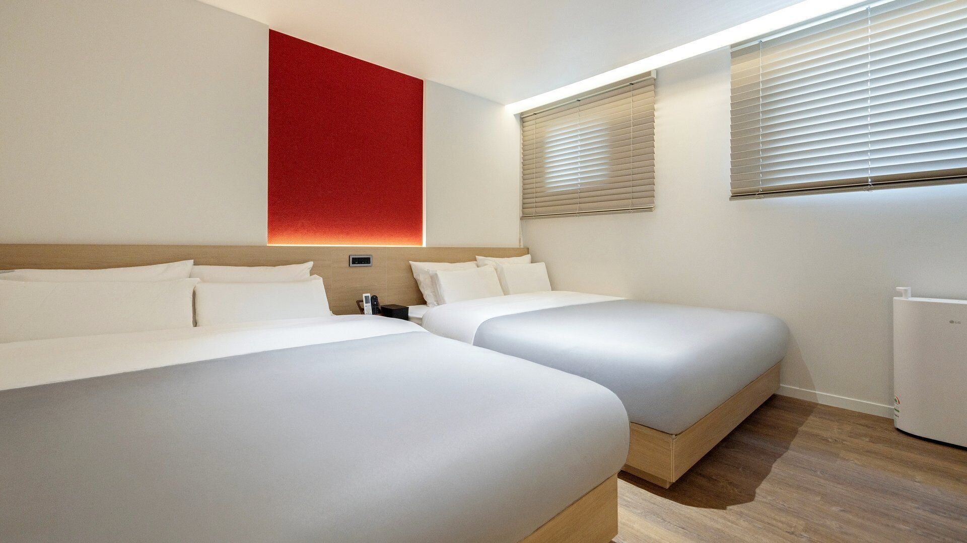 A hotel room with two beds, a red accent wall, and a window with blinds.