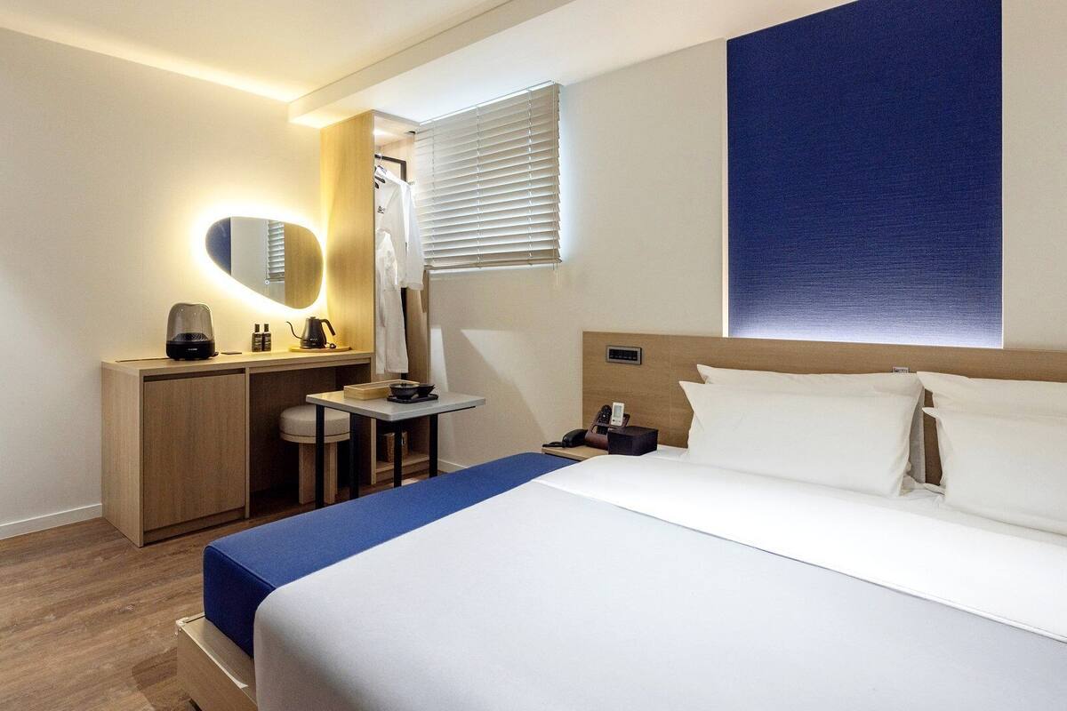 Habitación de hotel con cama, escritorio, espejo, ventana con persianas y una obra de arte colgada en la pared.