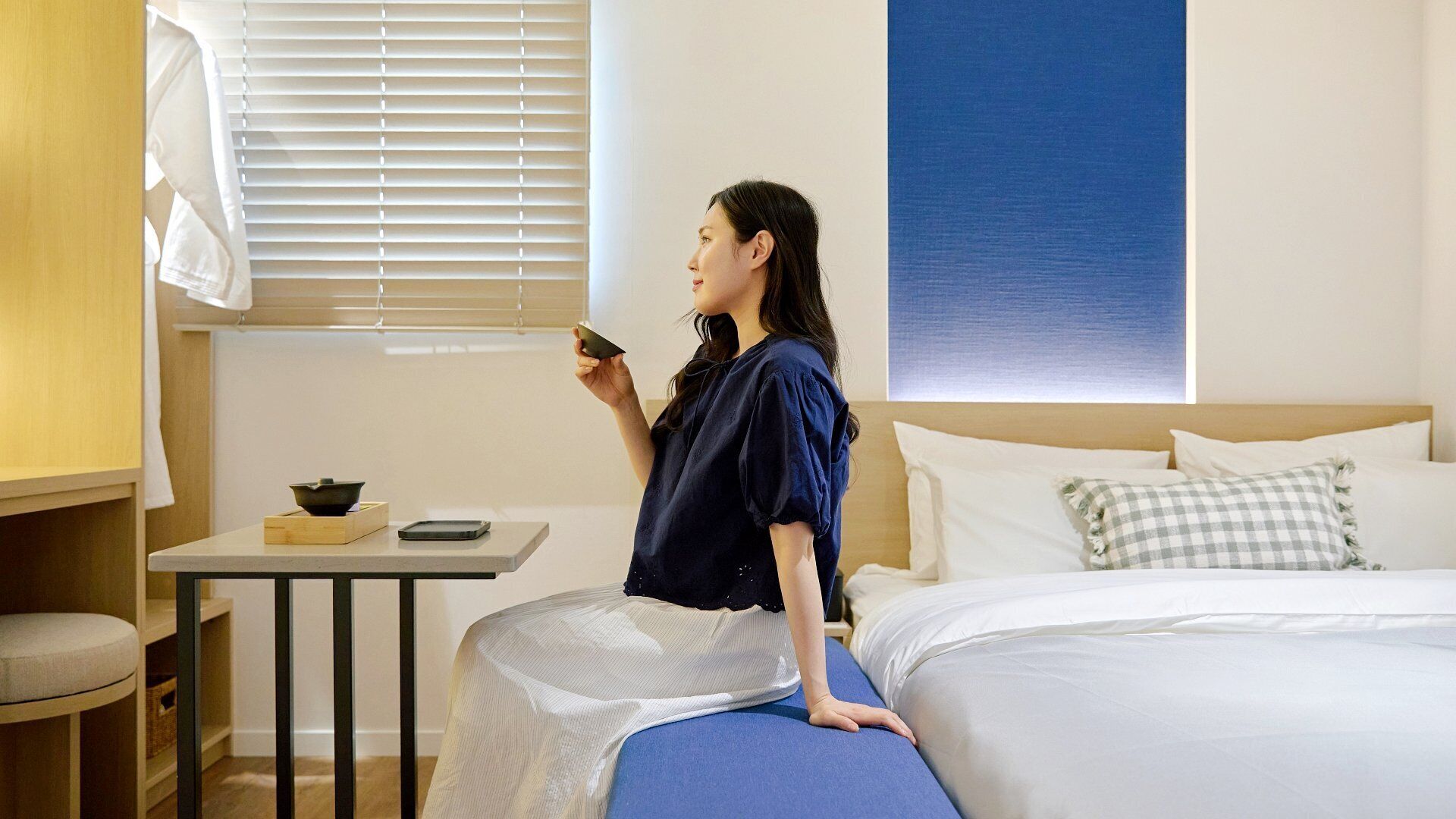 A room with a bed, a small table with a bowl and a smartphone, and a window with blinds.