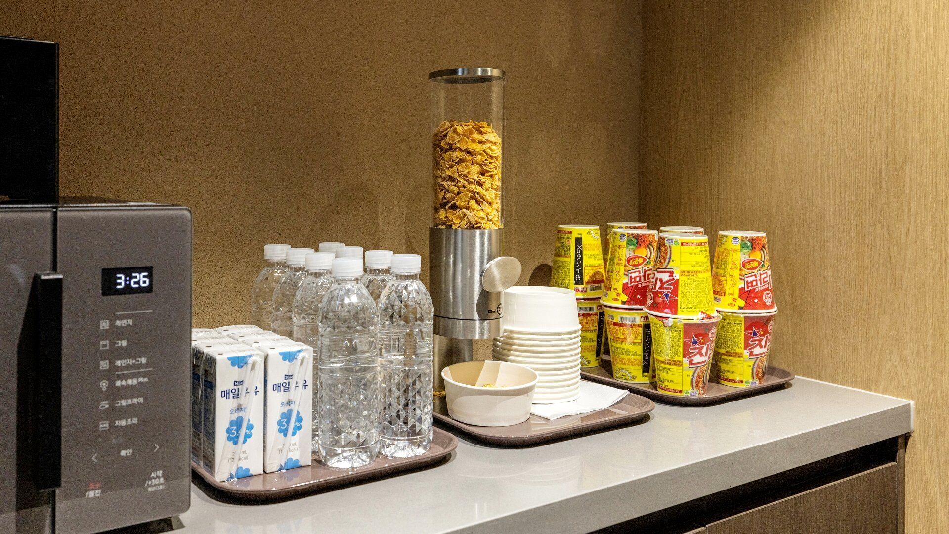 A breakfast buffet setup with a refrigerator, bottled water, milk cartons, cereal, and stacked cans.