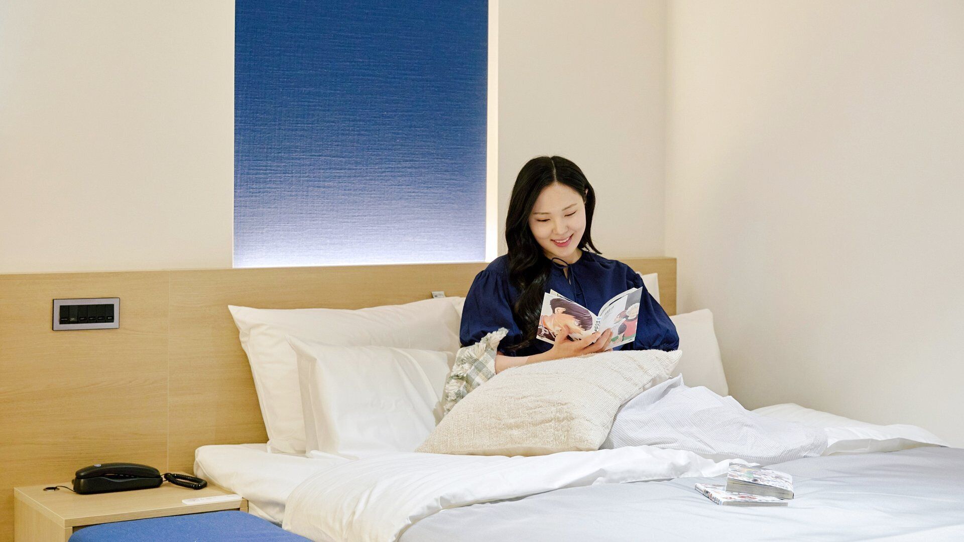 A hotel room with a bed, pillows, a bedside table with a phone, and a window with blinds.