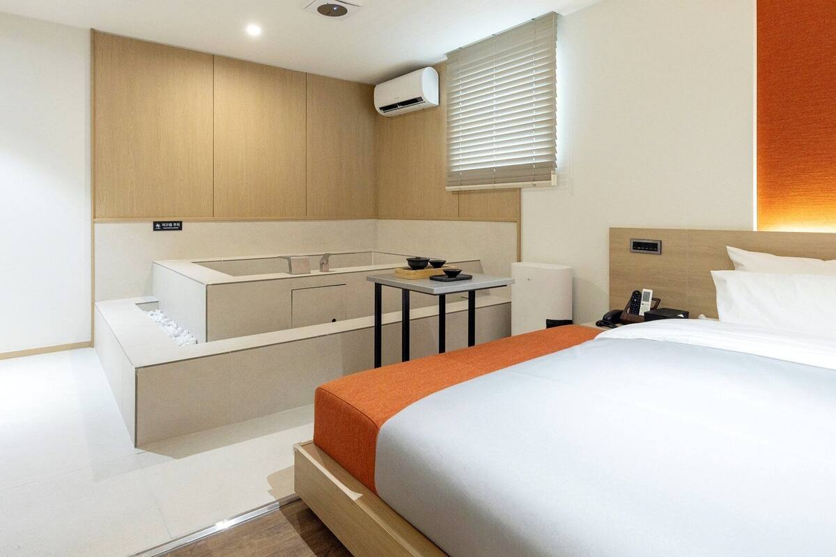Habitación de hotel con cama, una mesita, un banco empotrado, equipo de aire acondicionado y una ventana con persianas.