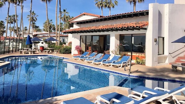 Une piscine entourée de chaises longues, de palmiers et d’un bâtiment doté d’un toit en tuiles.