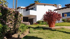 Une maison moderne à deux étages, avec une façade blanche, un toit en tuiles et un jardin bien entretenu, agrémenté de diverses plantes et d’un grand pot de fleurs rouge.