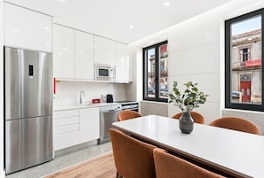 A modern kitchen with stainless steel appliances, a dining area with a table and chairs, and a view of the street through large windows. - GuestReady - Blossom Mosaic Daffodil Apartment (Porto)