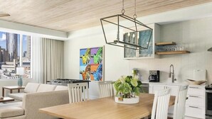 Un soggiorno moderno con un tavolo da pranzo in legno, sedie bianche, un divano e vista sulla città attraverso ampie finestre.