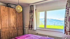 A bedroom with a bed, wooden wardrobe, a hanging light fixture, a window with a view of mountains, and a radiator. - Clair-Azur B 35 by Interhome (Crans-Montana)