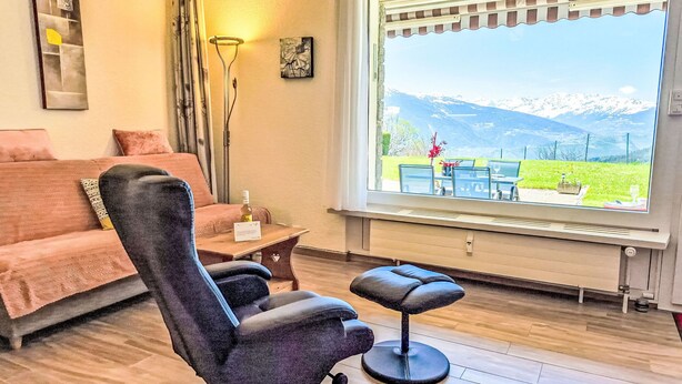 A room with a sofa, a massage chair, a table with a bottle, and a window with a view of mountains. - Clair-Azur B 35 by Interhome (Crans-Montana)