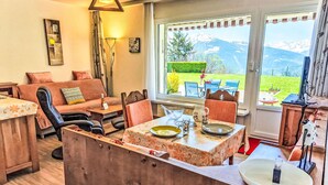 A cozy living room with a sofa, chairs, a dining table set for two, and a view of mountains through large windows. - Clair-Azur B 35 by Interhome (Crans-Montana)