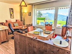 A cozy living room with a sofa, chairs, a dining table set for two, and a view of mountains through large windows. - Clair-Azur B 35 by Interhome (Crans-Montana)