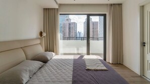 Une chambre moderne avec un grand lit, une vue sur de hauts immeubles et une porte-fenêtre coulissante.