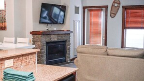 Ein Wohnzimmer mit Kamin aus Stein, einem darüber montierten Fernseher, einer Couch und einer Küchenzeile mit Arbeitsplatte und Geschirrspüler.
