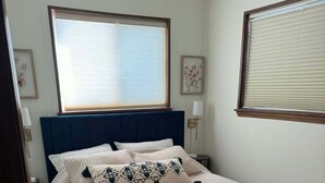 Uma cama bem arrumada com cabeceira azul, uma janela grande com persianas e uma luminária na parede.