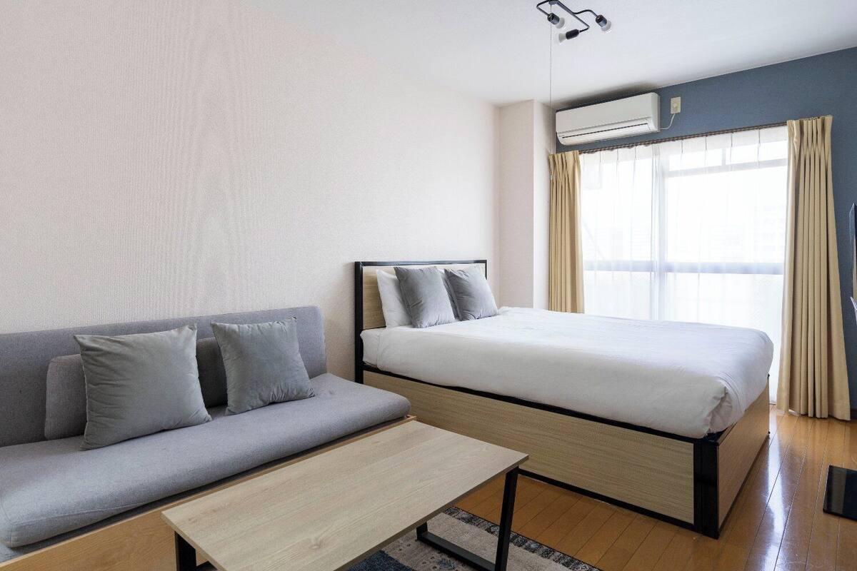 A hotel room with a bed, a sofa, a small table, and an air conditioning unit.