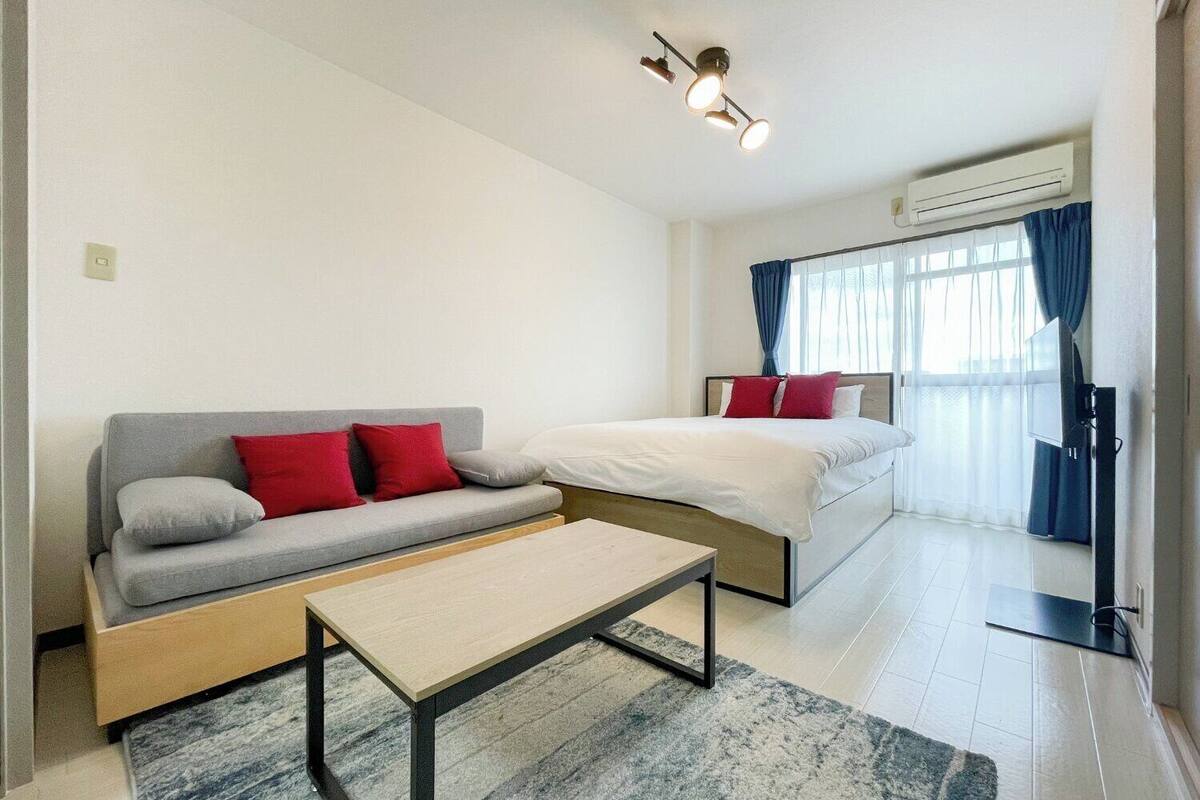 A modern hotel room with a grey sofa, a bed with red pillows, a wooden coffee table, and a large window with curtains.