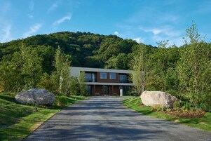 A modern two-story building with a paved driveway leading to a large entrance, surrounded by lush greenery and large rocks.