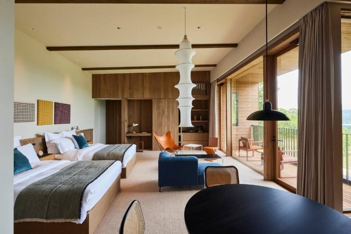 A modern hotel room with two beds, a dining area, a balcony with outdoor seating, and a large window.