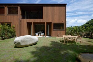A modern two-story building with a wooden exterior, a large rock in the foreground, and a dining area with outdoor furniture.