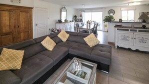A living room with a sectional sofa, a glass coffee table, and a kitchen area in the background. - Maison de charme conviviale (Montgivray)