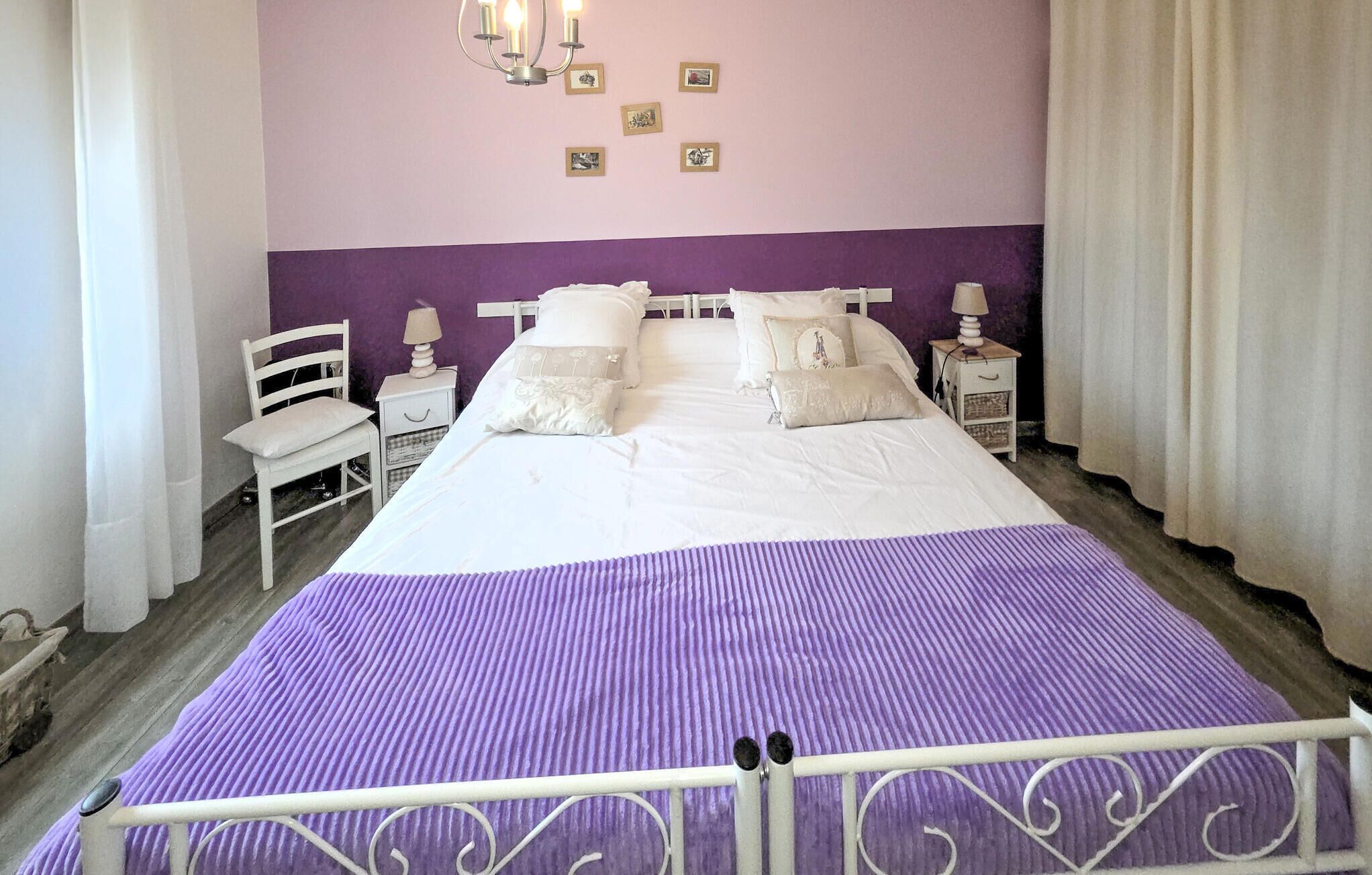 Una cama con colcha morada, ropa de cama blanca y cabecera de metal blanco.
