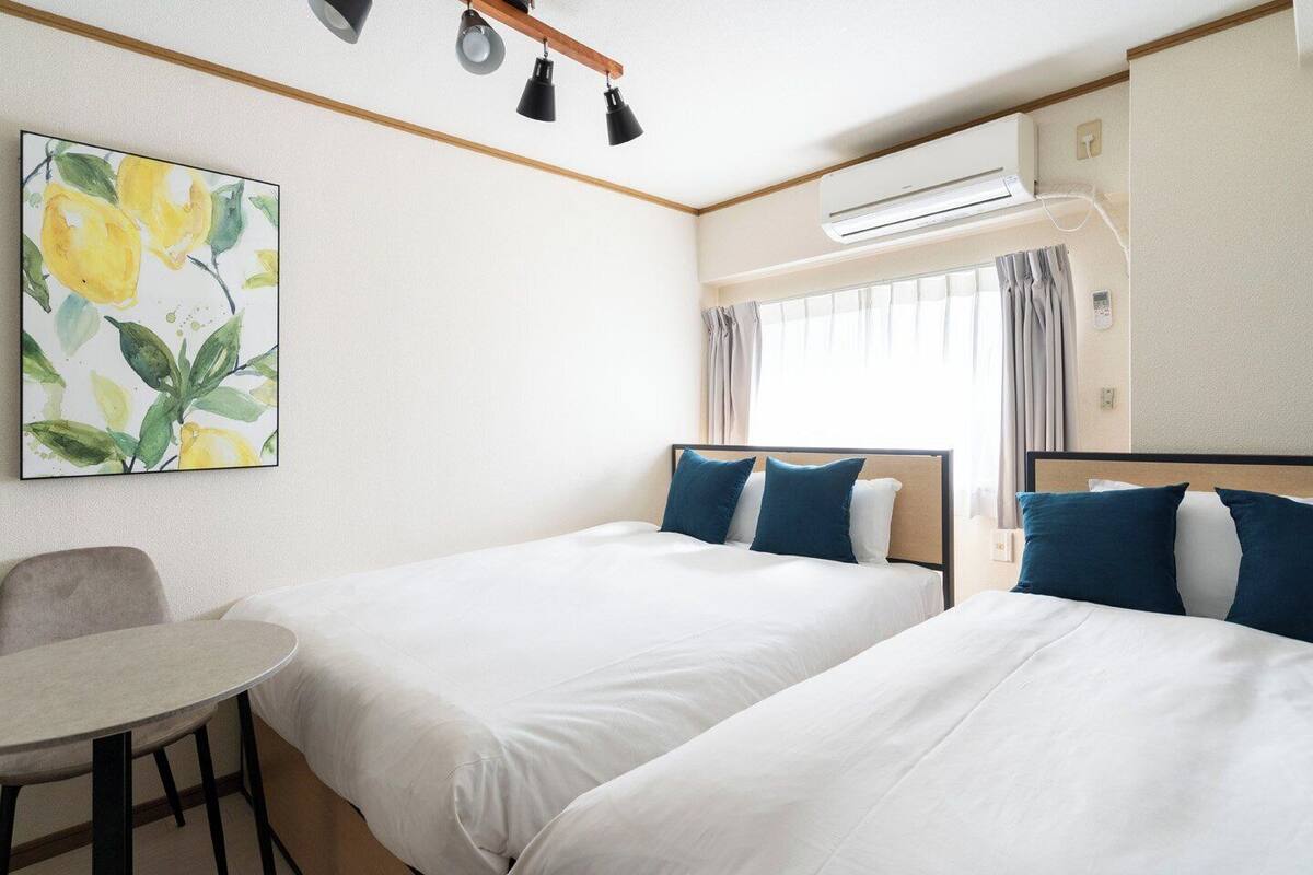 A hotel room with two beds, a table, a painting, and an air conditioning unit.