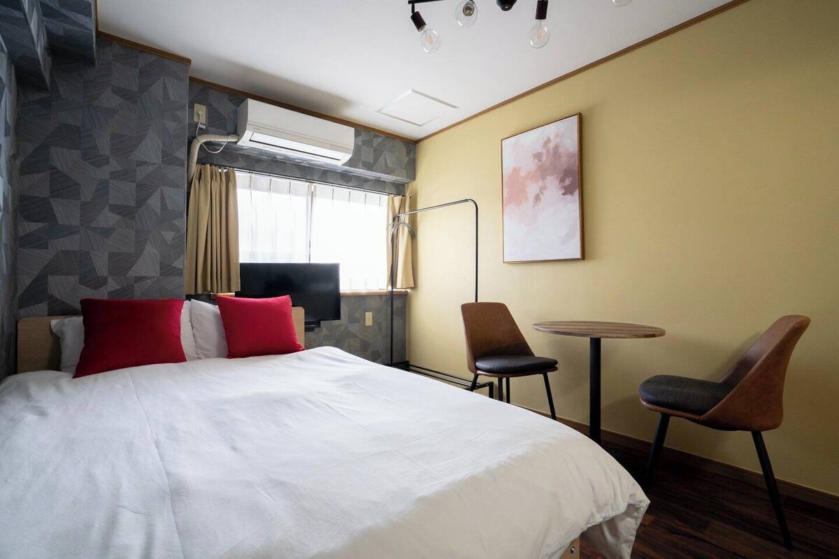 A hotel room with a bed, a desk, two chairs, an air conditioning unit, a television, and a painting on the wall.