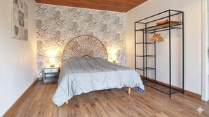 A bedroom with a wooden ceiling, a patterned wallpaper, a bed with a grey and white checkered bedspread, two bedside lamps, and a black metal shelf unit with clothes. - Cozy home in Saint-Christophe-du-Foc with WiFi (Saint-Christophe-du-Foc)