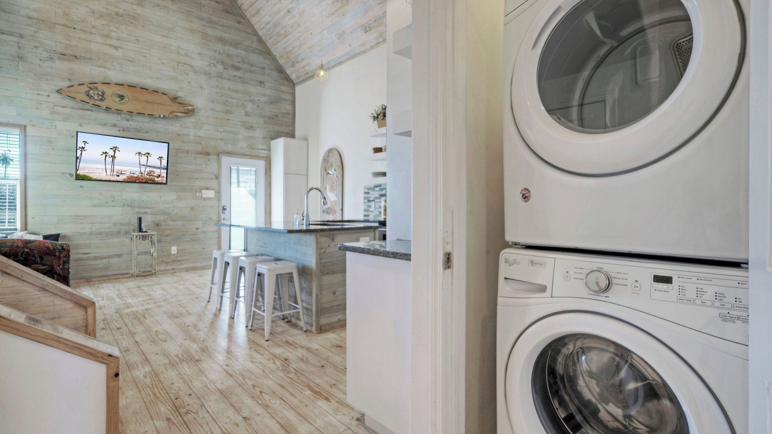 A laundry room with stacked washers and dryers, a kitchen area with a bar, and a living space with a large TV and wooden flooring.
