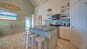 Una cocina con techo de madera, una isla grande con taburetes, armarios blancos y una vista del océano en la pared.