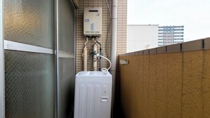 A water heater mounted on a tiled wall with a metal vent pipe, a metal fence, and a building in the background. - Mansion Castle (Osaka)