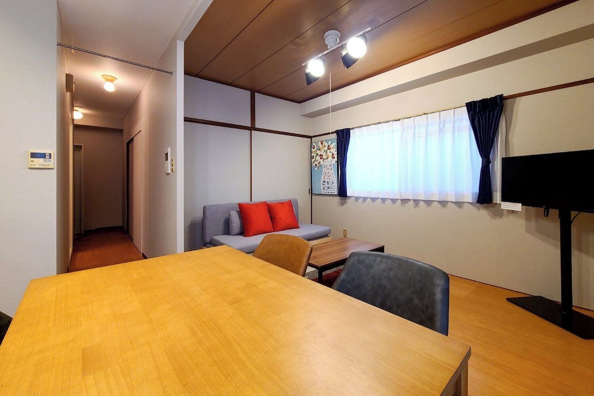 A room with a wooden table, a sofa with red cushions, a television, and a window with curtains.