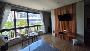 A modern living room with a large window, a flat-screen TV mounted on a wooden panel wall, a small round table, two chairs, and a wooden floor. - Peya Rama-7 (Bang Kruai)