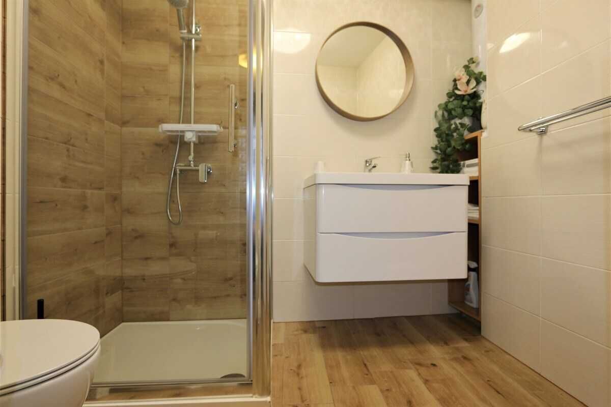 A bathroom with a glass-enclosed shower, a white sink, a mirror, and a wooden floor.