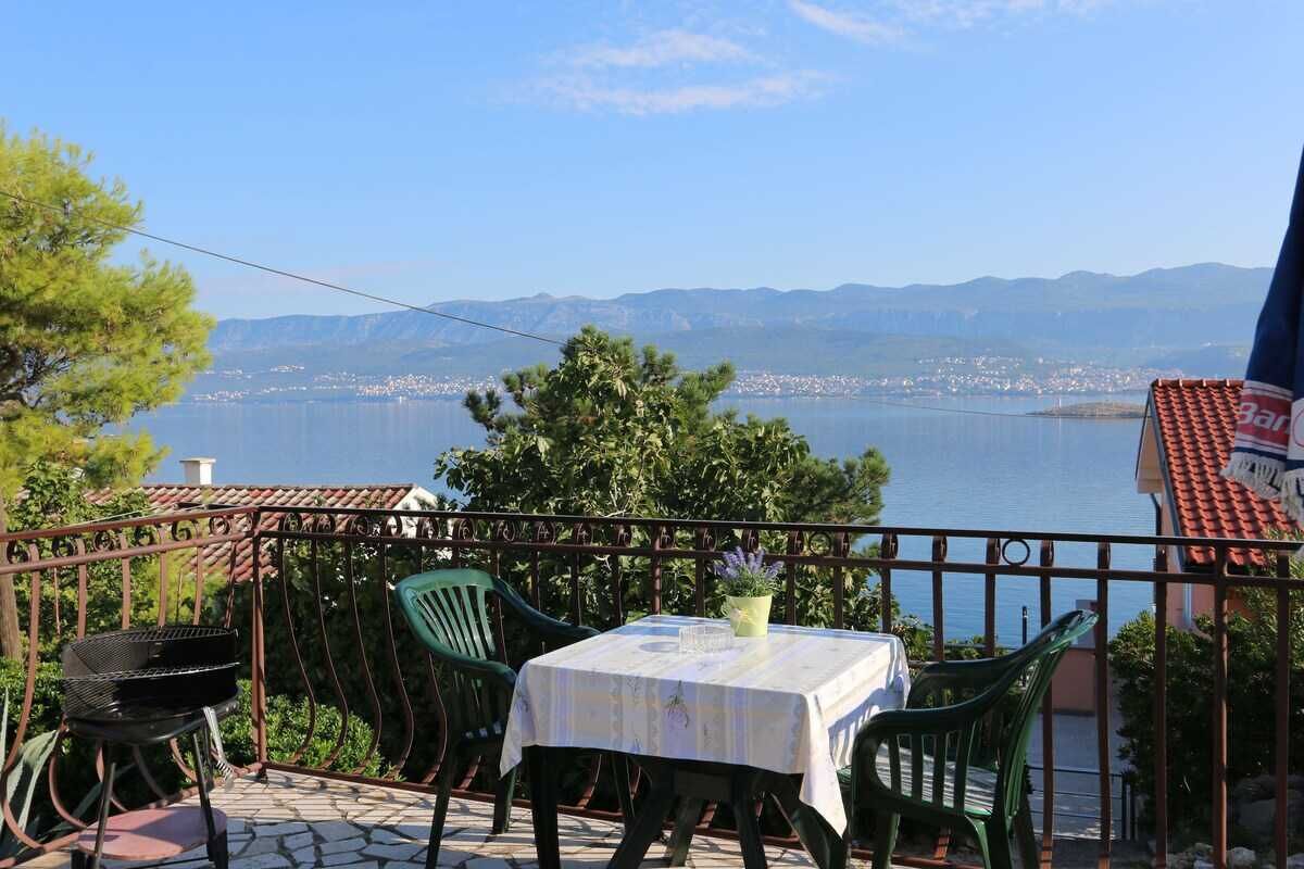 Une terrasse dotée d’une table, de chaises et d’un barbecue, offrant une vue sur une étendue d’eau et sur des montagnes lointaines.
