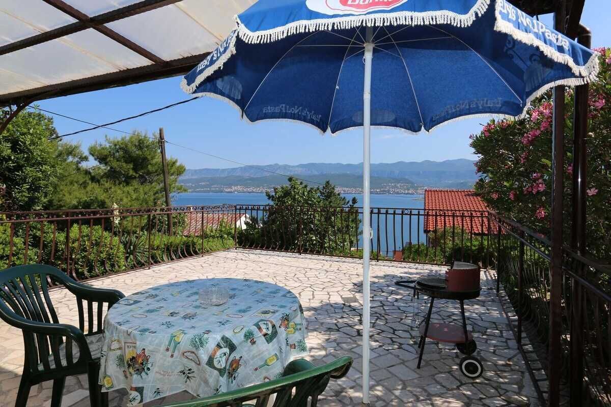 A covered outdoor seating area with a table and chairs, a blue umbrella, and a view of the sea and mountains.