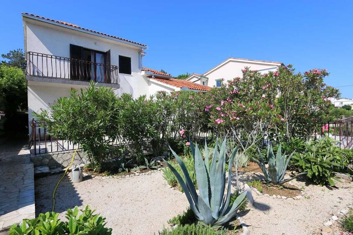 Un bâtiment à deux étages doté d’un balcon, entouré d’un jardin où poussent différentes plantes et fleurs.