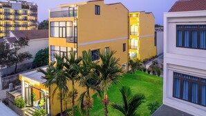 A multi-story building with a yellow facade, a visible entrance, and a small garden area with palm trees. - Corner Centre Villa Hoi An (Da Nang)