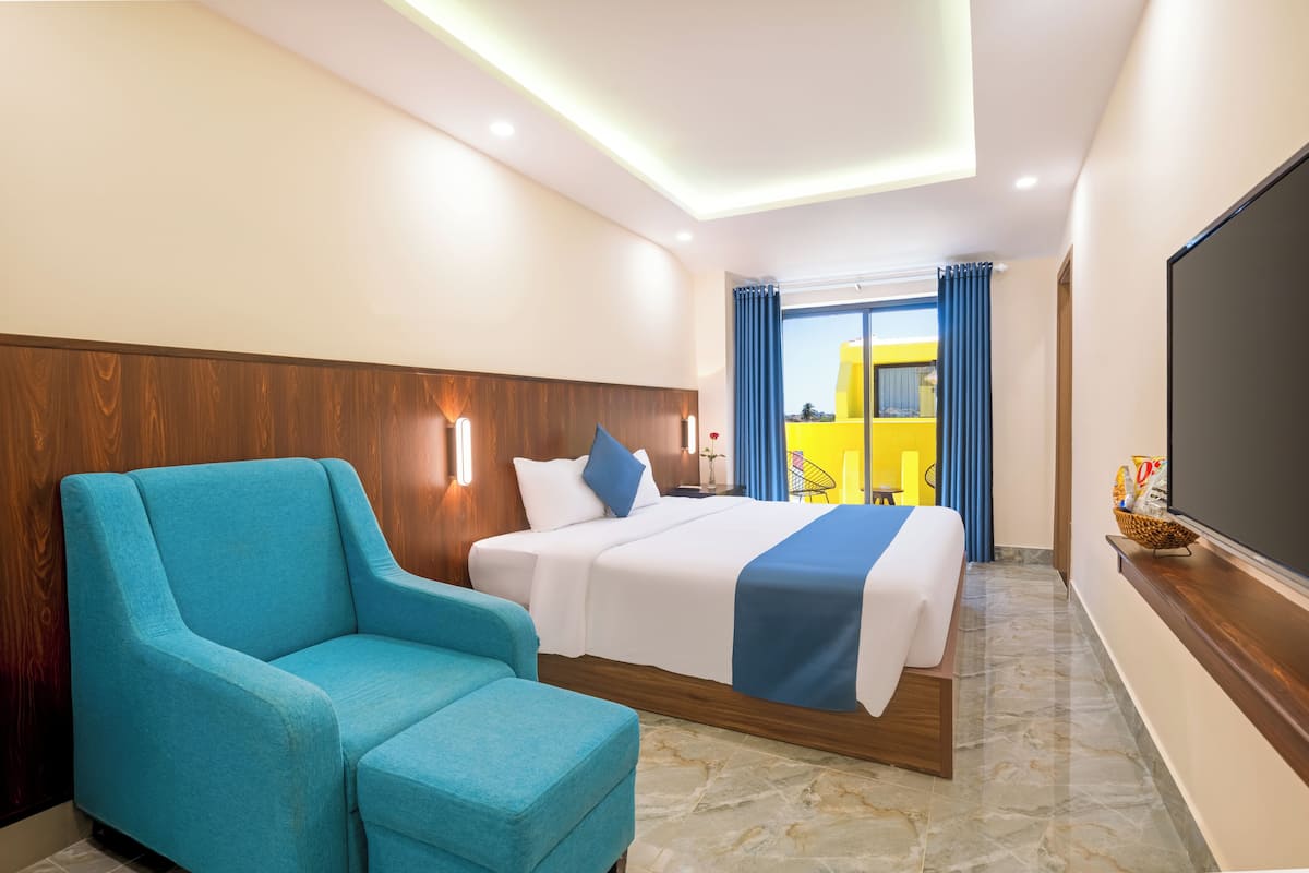 Habitación de hotel con una cama, un sillón azul, televisión y vista a un edificio amarillo.