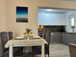 A dining area with a table set for two, a kitchen with white cabinets, and a painting on the wall.