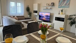 Uma sala de estar com sofá, televisão, mesa de jantar com pratos e copos, e um aparador com uma planta.