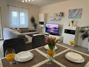 A living room with a sofa, TV, dining table with plates and glasses, and a TV stand with a plant. - Apartment Maticic gore (Šilo)