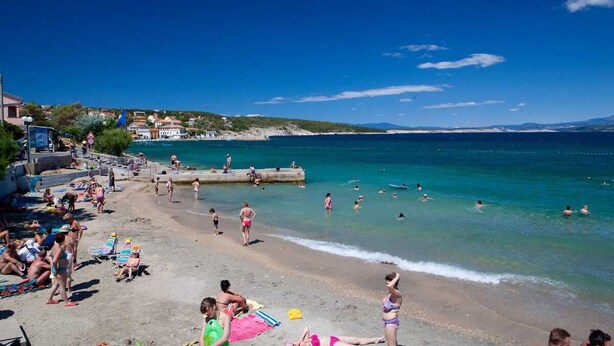 Uma praia com pessoas nadando e tomando sol, um píer e edifícios ao fundo.
