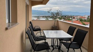 Uma varanda com mesa e cadeiras, um vaso de planta e vista para uma cidade e montanhas.