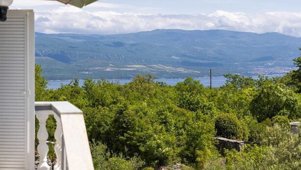 A view of a mountainous landscape with a body of water in the distance, a white building with a balcony, and dense green foliage. - Apartment Maria Zestilac (Žestilac)