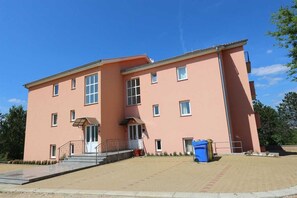 Un edificio de varios pisos con techo plano, una entrada central con escalones y un bote de basura azul.